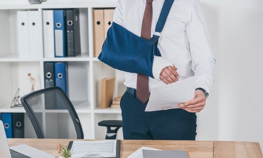 Injured worker with broken arm in sling holding workers' compensation settlement document at desk